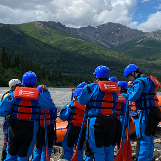 Group of rafters in blue dry suits and orange life jackets preparing for a Nenana River rafting trip in Denali National Park, Alaska, with mountain views in the background.
