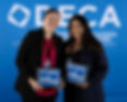 Two young women in business attire smile in front of a blue background that reads DECA. 
