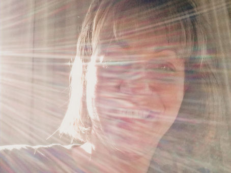 A smiling woman bathed in dramatic rays of light, wearing a shirt with a heart graphic, representing spiritual illumination and openness to unexpected guidance.