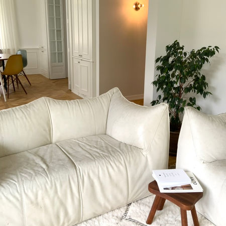 helles weisses Wohnzimmer mit beigem Vintageledersofa, beigem Beniteppich, Holzhocker, im Hintergrund Pflanze und Blick ins Esszimmer mit Wandlampe; Dekor, Homedekor, Styling, Homestyling