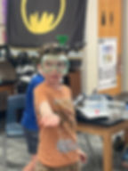 A student in a Drone Legends classroom wears safety goggles and a head-mounted drone launcher while participating in a hands-on STEM activity. The room features themed decor and Drone Legends equipment, fostering fun and engaging drone-based learning. Learn more at dronelegends.com.