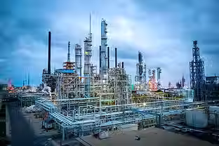 Wide-angle photo of an industrial refinery at dusk, with tall process towers, piping, and illuminated structures under a cloudy sky.