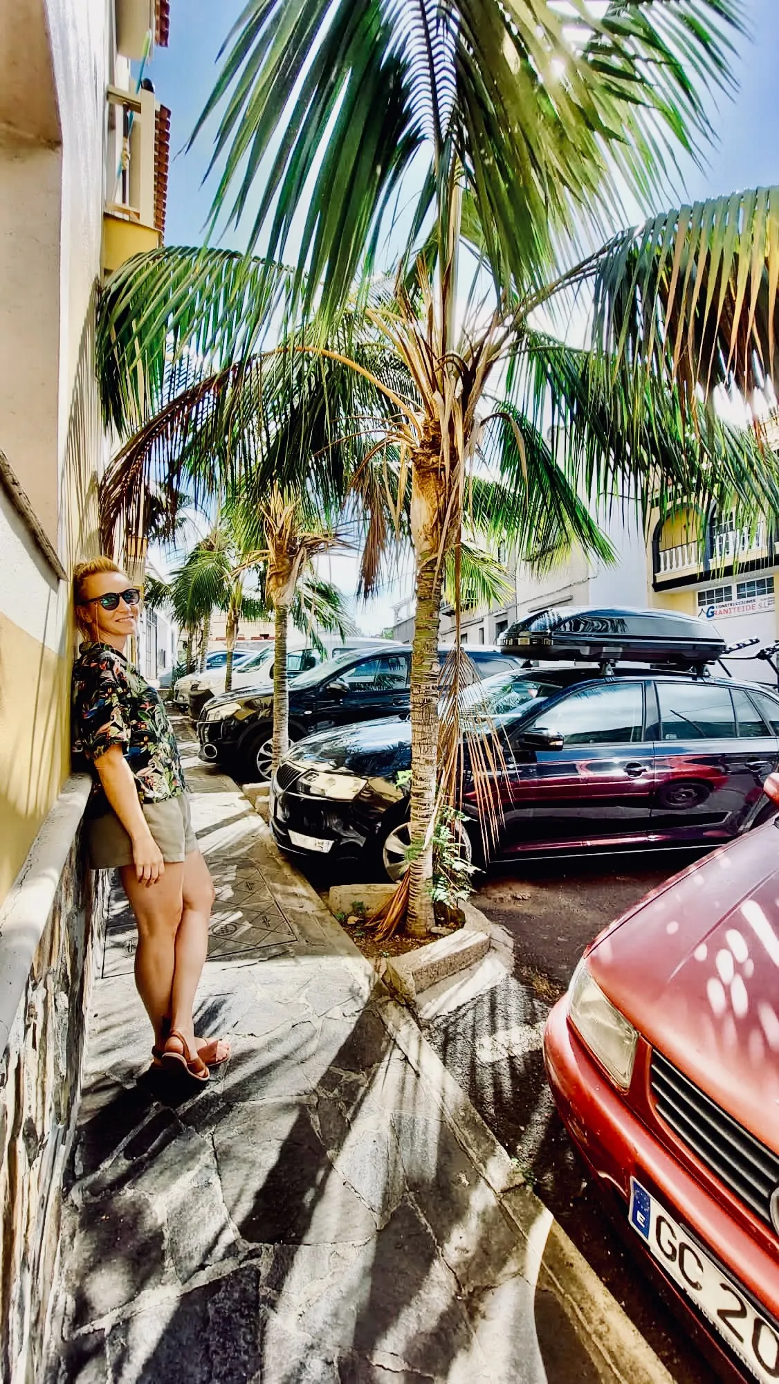 Spacer wśród palm na północno-zachodniej Teneryfie – codzienne życie na wyspie, słońce, cienie liści i zaparkowane samochody przy wąskiej uliczce.