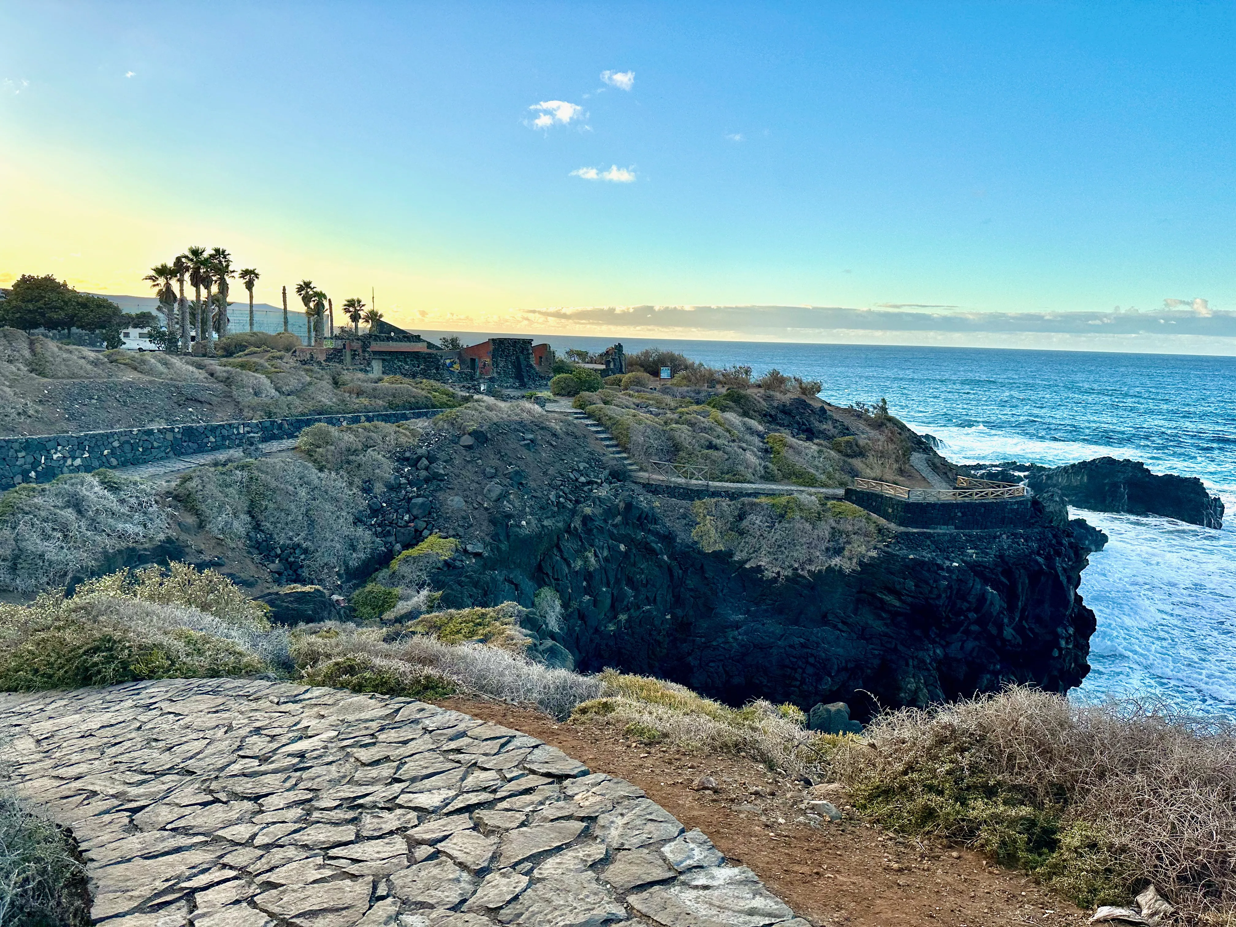 Kamienny chodnik biegnący nad wulkanicznymi klifami, z widokiem na Atlantyk i niską zabudowę na horyzoncie, o zachodzie słońca.