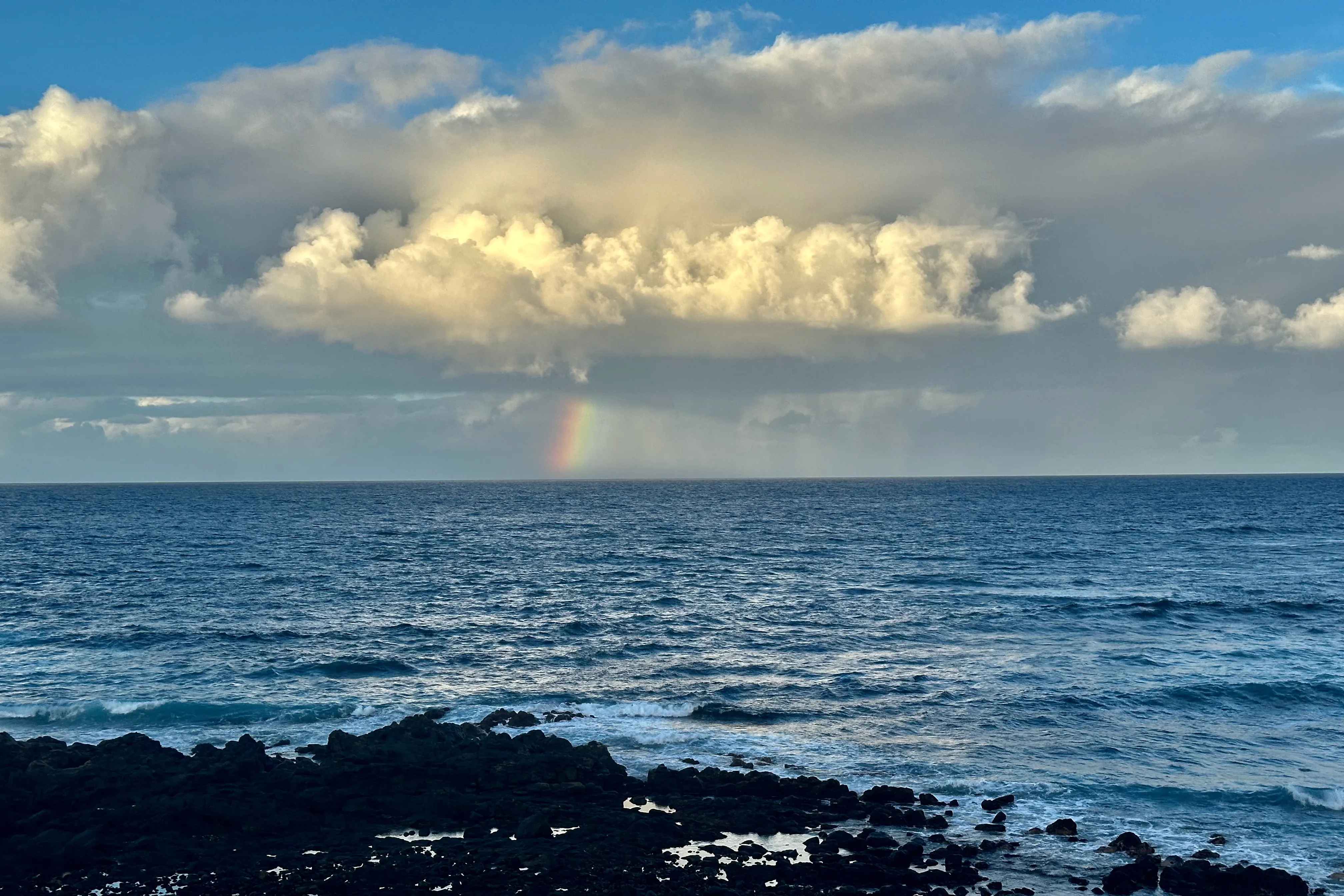 Tęcza schodząca do Atlantyku pod masywną chmurą, widok z kamienistego wybrzeża północnej Teneryfy.