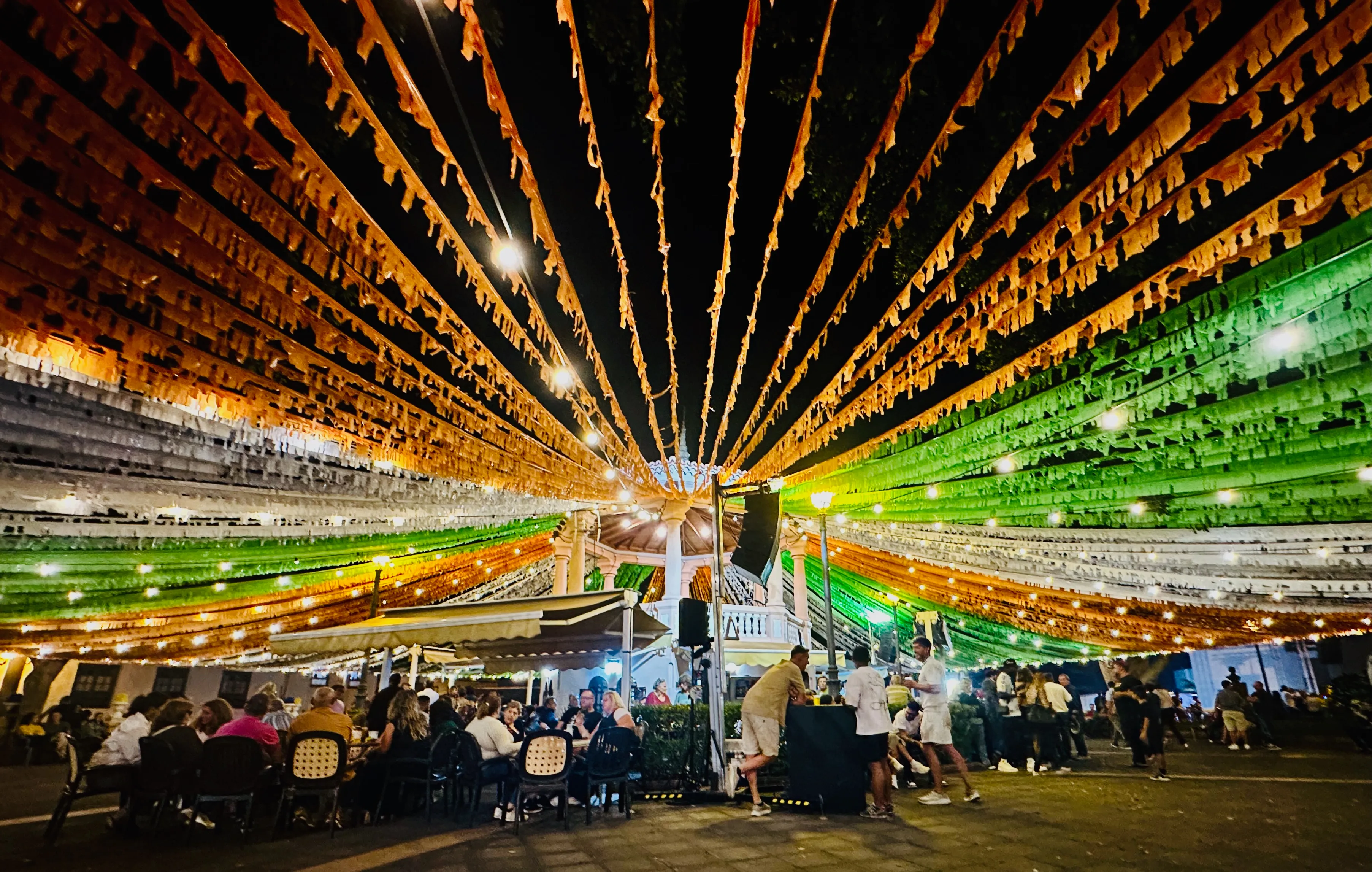Nocna fiesta na głównym placu miasteczka na Teneryfie – kolorowe girlandy nad głowami, światła i mieszkańcy siedzący przy stolikach na świeżym powietrzu.
