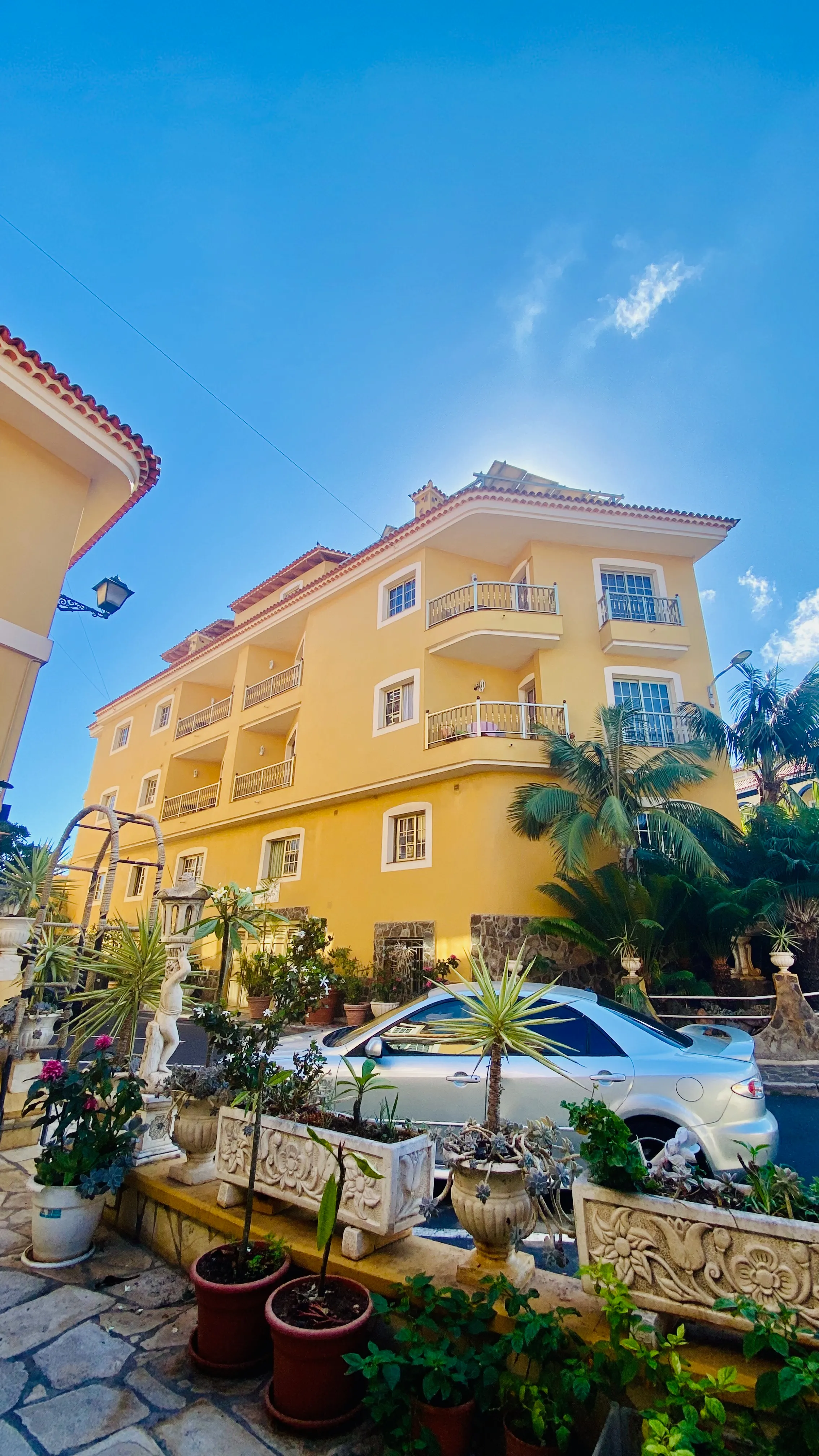 Żółty kanaryjski apartamentowiec z balkonami na tle błękitnego nieba – architektura północnej Teneryfy i rośliny w donicach przed domem