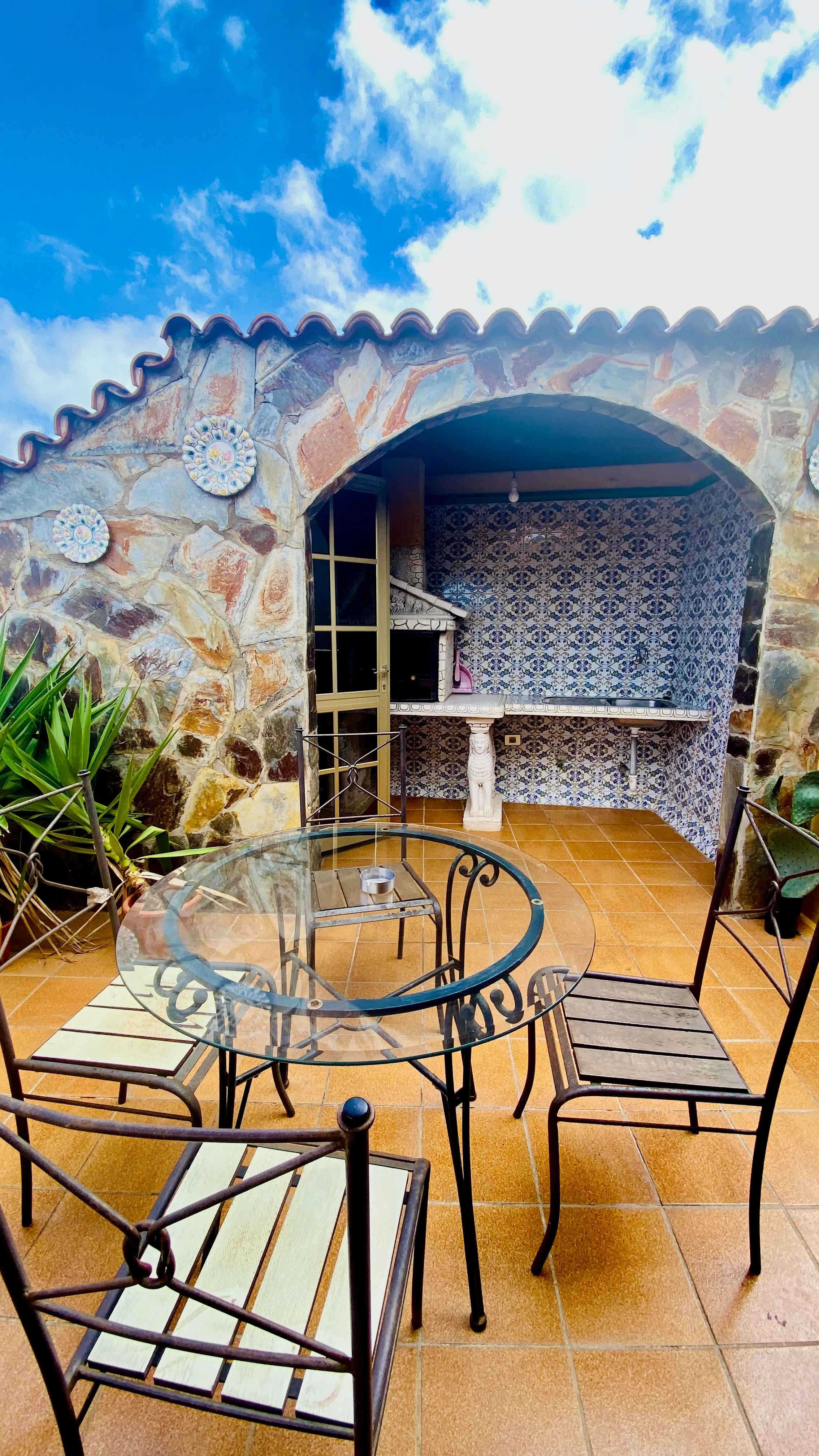 Przytulny kanaryjski taras na Teneryfie – kamienny łuk, kafelki w tradycyjne wzory, grill i szklany stół idealny na leniwe popołudnia.
