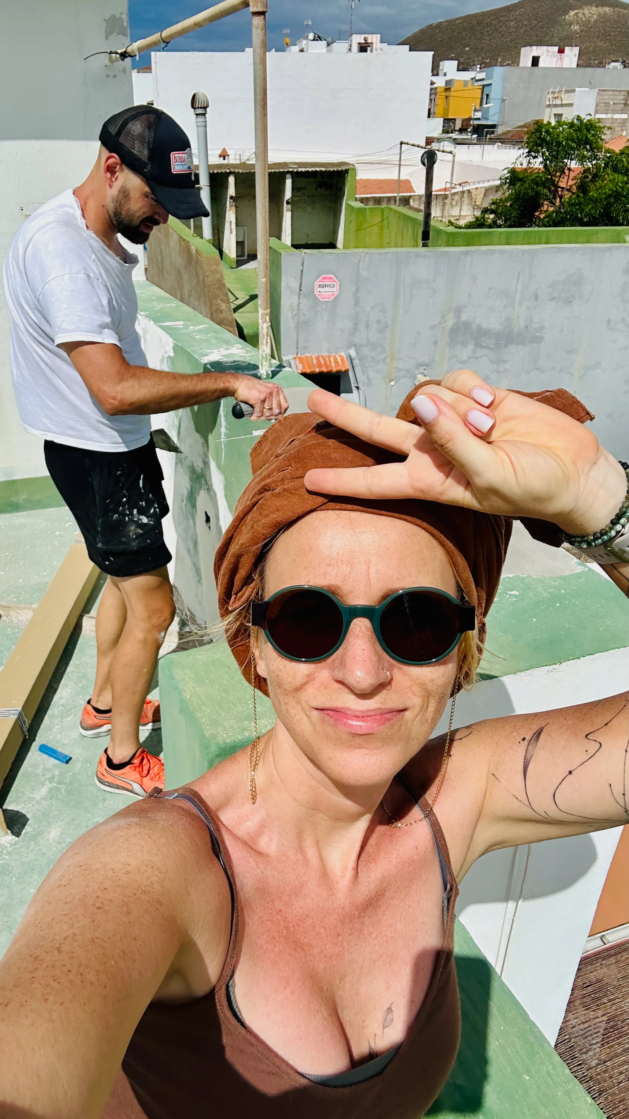 Remont dachu w kanaryjskim miasteczku na Teneryfie – para pracuje w słońcu na tarasie, z kolorową zabudową i wzgórzami w tle.