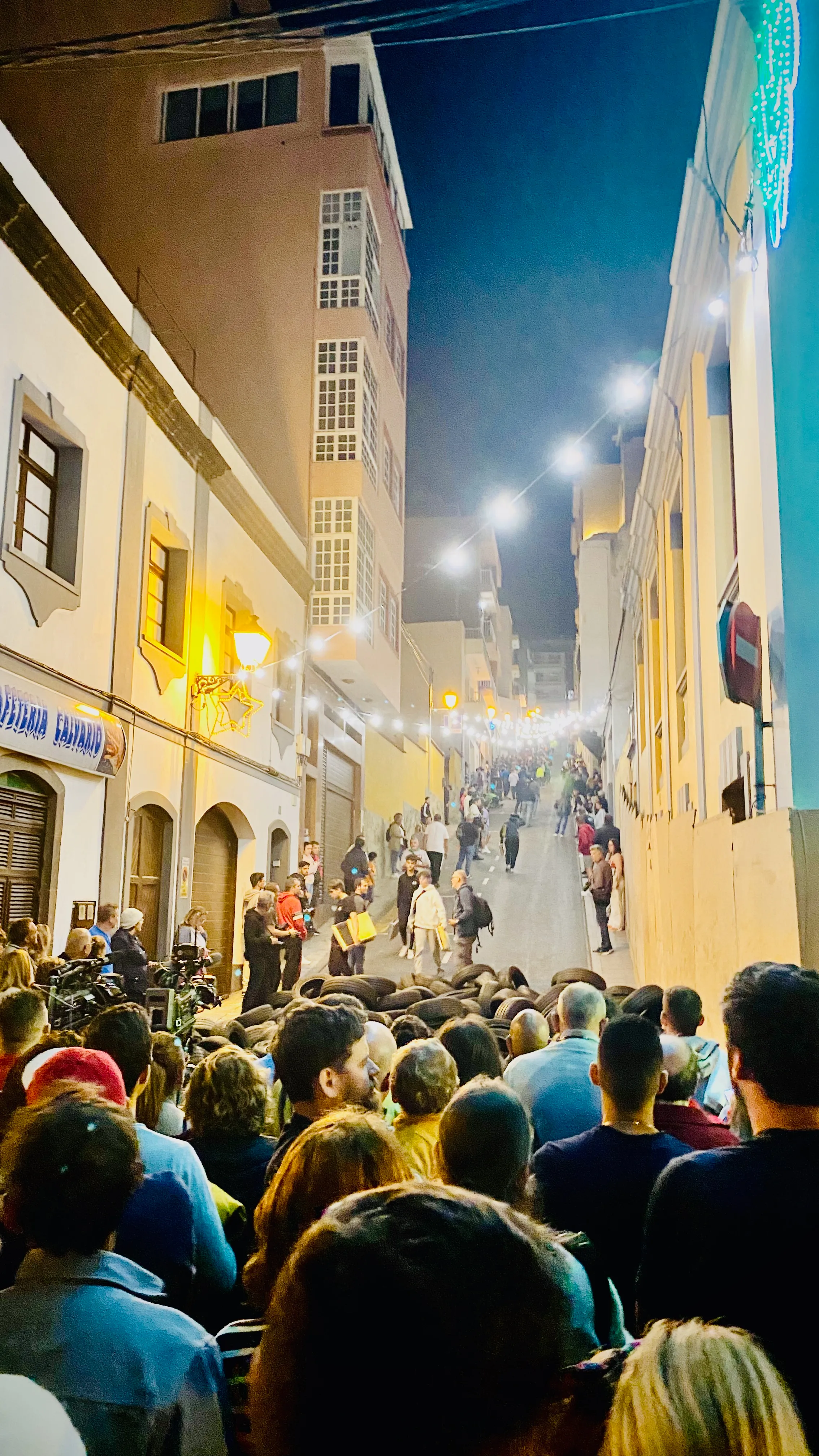 Tłum ludzi na stromej ulicy podczas lokalnej fiesty na Teneryfie, z oponami zabezpieczającymi zjazdy i rozświetlonymi kamienicami po obu stronach.