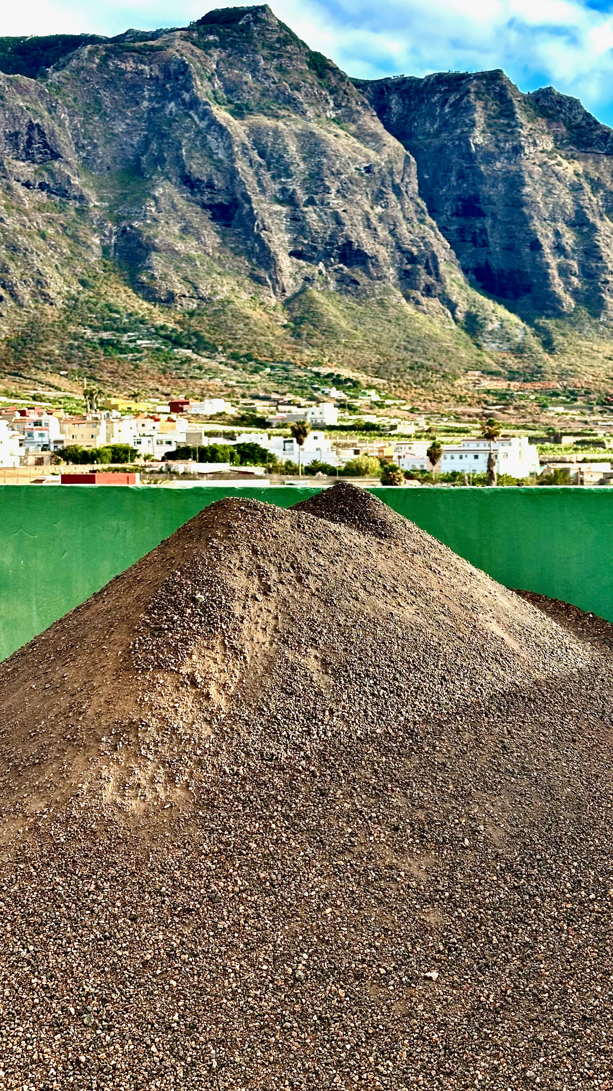 Stos wulkanicznego żwiru na tle zielonej doliny i stromych klifów północno-zachodniej Teneryfy, z białymi domami u podnóża gór.