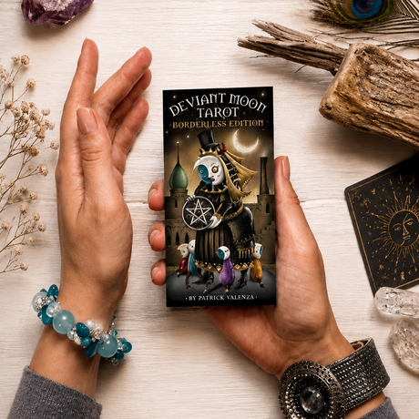 Manos sostienen una baraja de tarot Deviant Moon Tarot Borderless Edition; mesa ritual con cuarzo, pluma, madera y otra carta negra.
