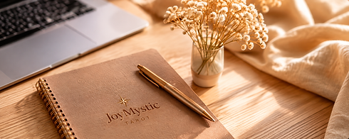 Cuaderno de JoyMystic Tarot y bolígrafo dorado sobre un escritorio de madera en tonos cálidos.