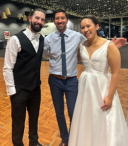 Three people pose and smile at a wedding reception, where string lights twinkle overhead. The bride wears a white gown, the groom dons a black vest and bow tie, and a guest stands between them—ready to dance thanks to the talented wedding DJ.