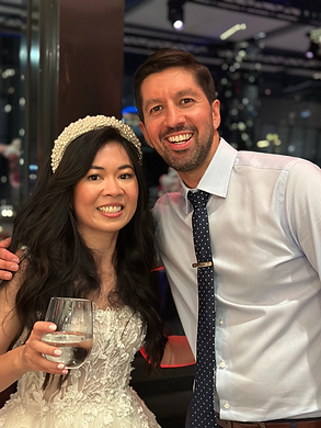 A smiling bride in a white lace dress holding a drink stands next to a man in a polka dot tie. They’re posing together indoors at an event, capturing the joy and energy often seen at Melbourne weddings with great rhythm and beats.