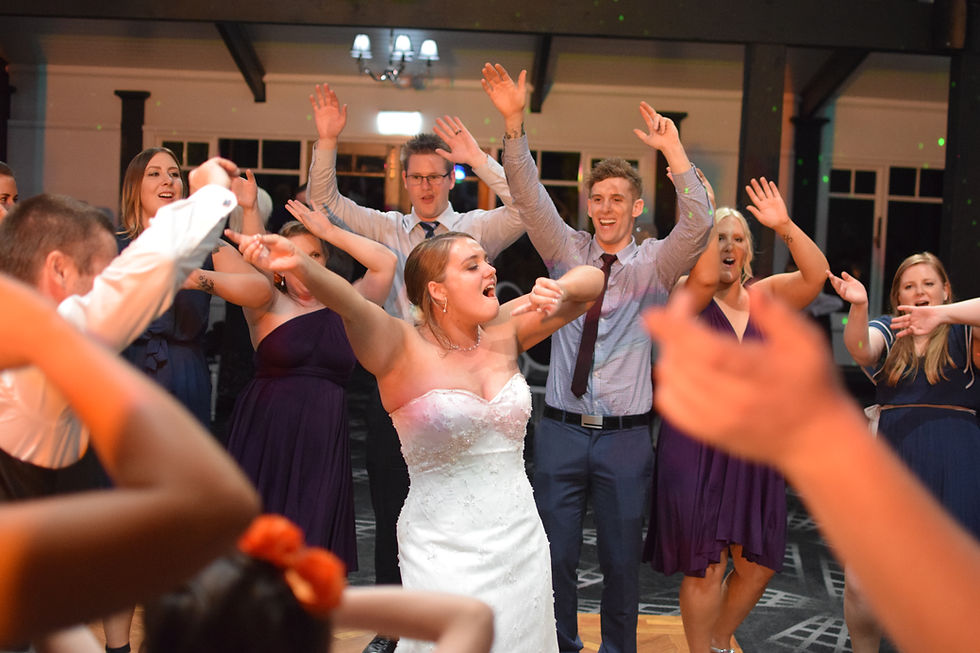A bride in a white gown joyfully sings and dances with friends and guests, all raising their arms and smiling during a lively wedding celebration indoors with an energetic wedding DJ Melbourne.