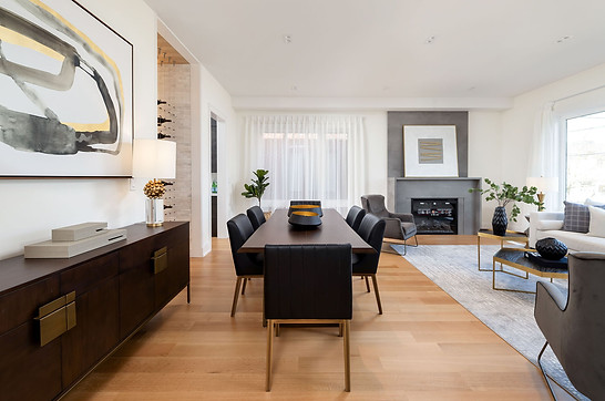 Toronto custom home design build project by Nestwood Home, Powered by Z Square Group. The image depicts a spacious living room with a dining table, a fireplace, and several pieces of furniture such as a couch, a chair, and a coffee table. There is also a large painting hanging on the wall above the fireplace. The room is well-lit and has a wooden floor.