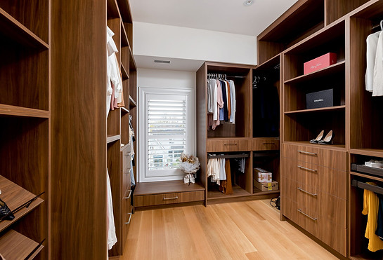 Toronto custom home design build project by Nestwood Home, Powered by Z Square Group. The image depicts a walk-in closet with wooden shelves and drawers, a white window, and a wooden floor. The closet is well-organized with various clothing items, shoes, and accessories displayed on the shelves and in the drawers.