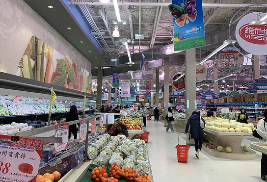 Z Square Group. The image depicts a busy supermarket with a variety of fruits and vegetables on display. Customers can be seen shopping for their groceries, and there are several signs and banners hanging from the ceiling, showcasing different products and promotions.