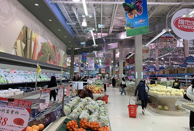 Z Square Group. The image depicts a busy supermarket with a variety of fruits and vegetables on display. Customers can be seen shopping for their groceries, and there are several signs and banners hanging from the ceiling, showcasing different products and promotions.
