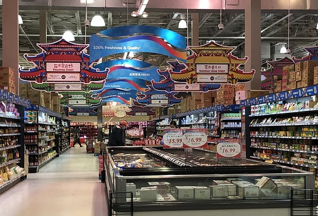 Z Square Group. The image shows a well-organized and visually appealing grocery store with various products displayed on shelves and in bins. There are colorful signs and banners hanging from the ceiling, promoting the store's products and services.