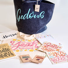 Bild mit verschiedenen selbstdesignten Produkten, es zeigt verschiedene beschriftete Taschen und Mäppchen, Karten, Pins und ein Notizbuch
