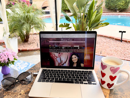 Laptop open to yomary.com with a cup of coffee with pink, red & burgundy hearts on the right, sunglasses on left and a pool & palm trees in the background.