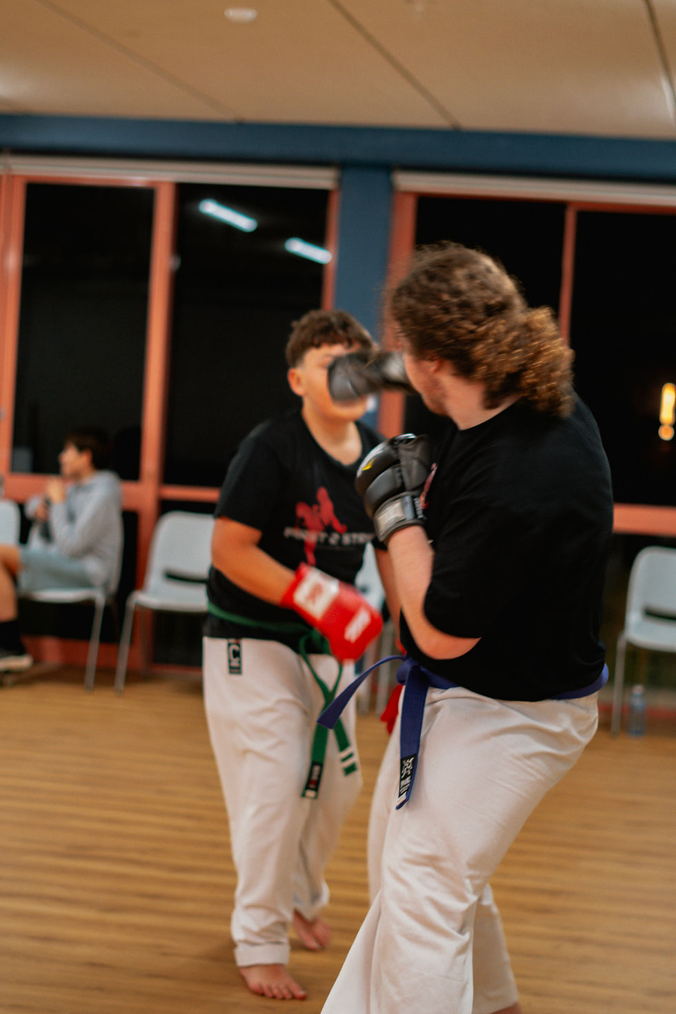 Two martial arts sparring partners in a gym trade punches, wearing gloves and white pants with colored belts.