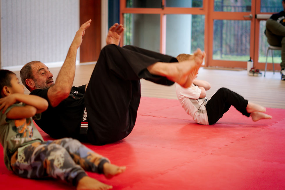 Three people, including a man and two children, perform sit-ups on a red mat in a gym. The setting is bright, with large windows.