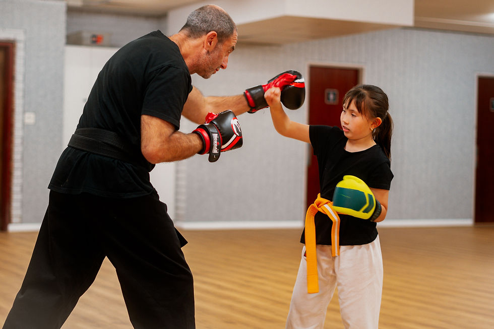 Man and girl practice boxing in a gym. The man holds focus mitts, and the girl has a yellow belt. Both wear black shirts.
