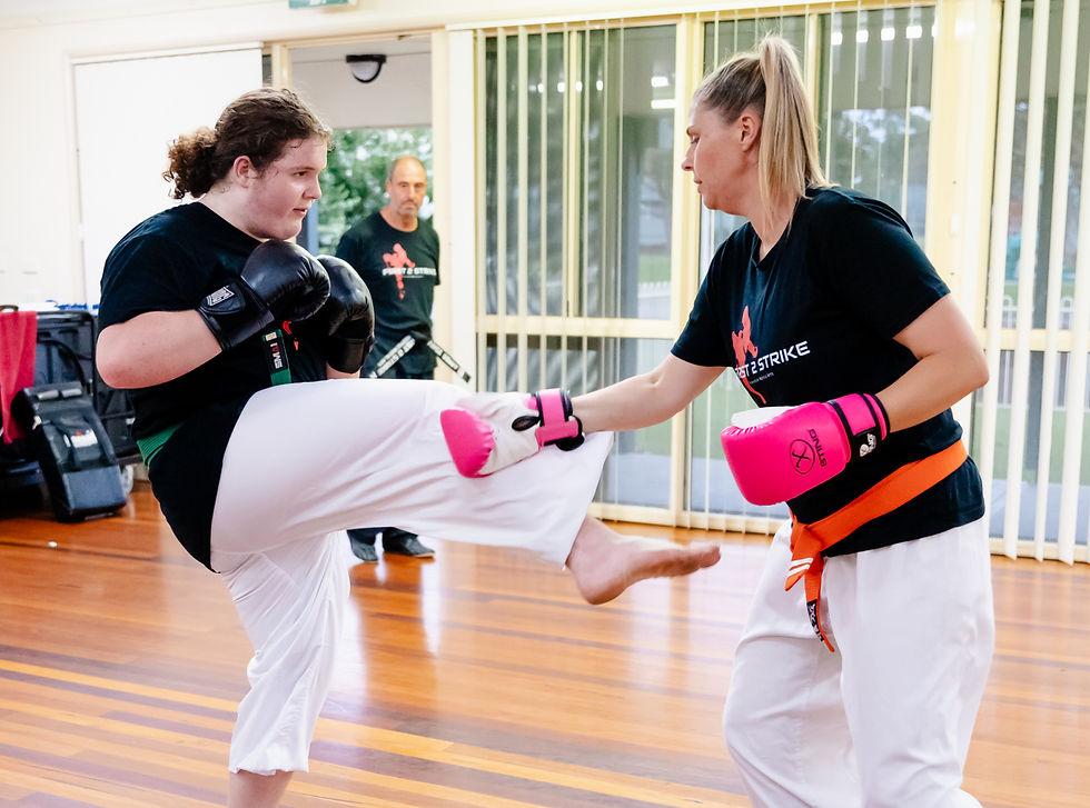 Two women spar in a bright dojo, one blocking a kick while a man watches; both wear black shirts, white pants, and pink gloves.