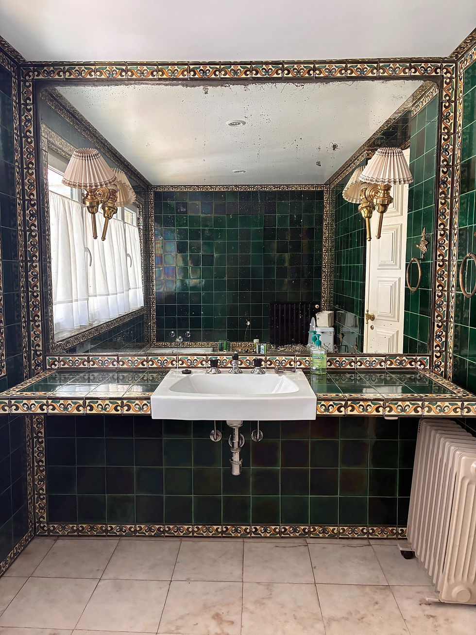 Green tiles bathroom sink and wall with a centered rectangular mirrow in the center and golden colour patterns as frame.