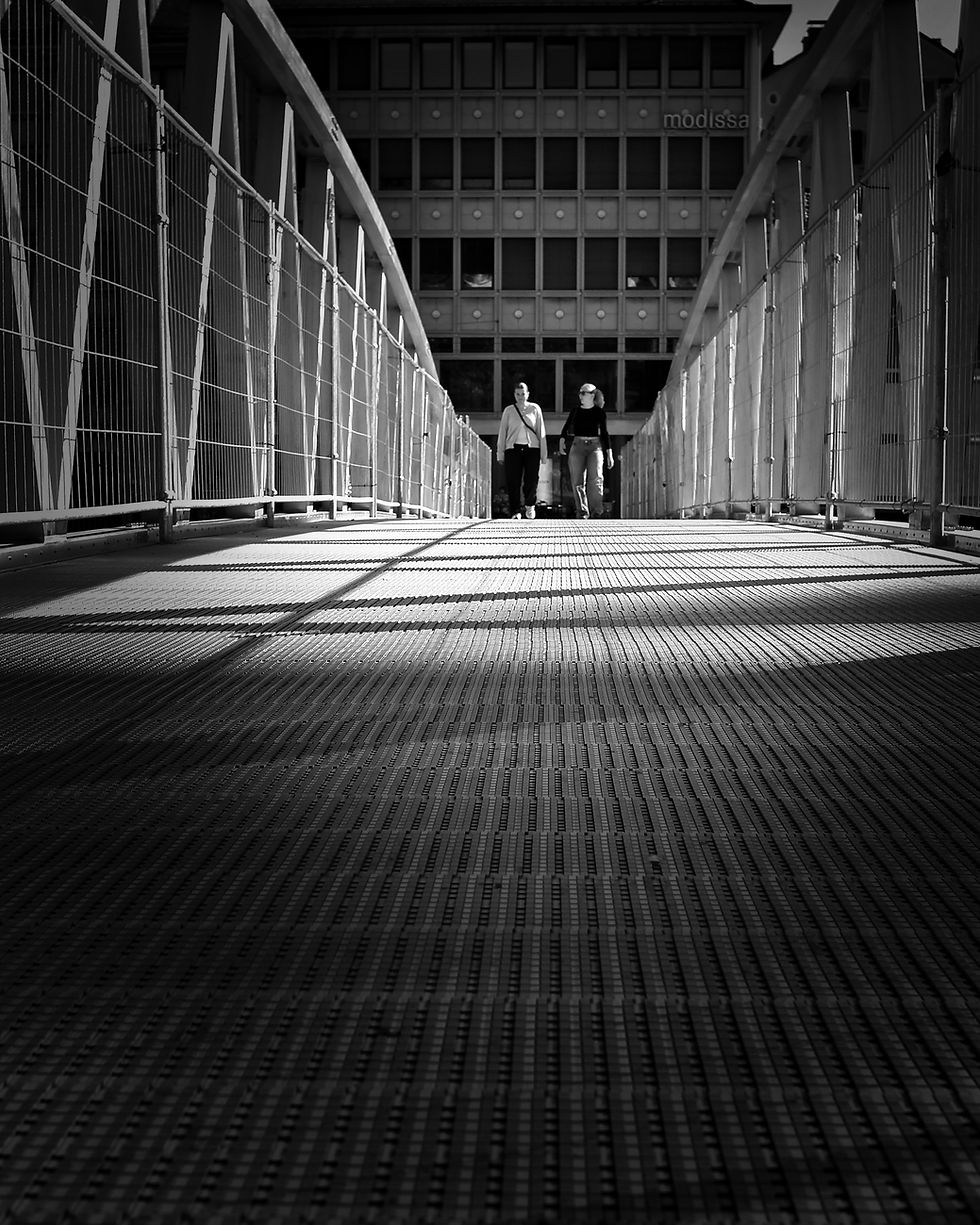 Black and white photograph of a metal footbridge with geometric railings and long shadows.