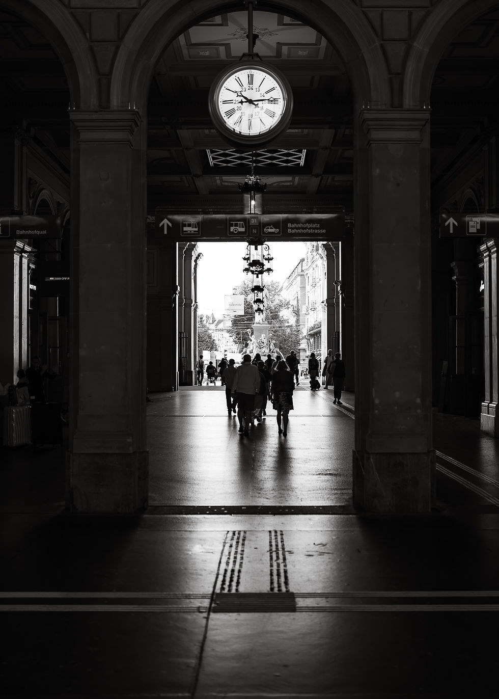 Black and white photograph of a railway station passage with a large clock and people in the bright passageway.