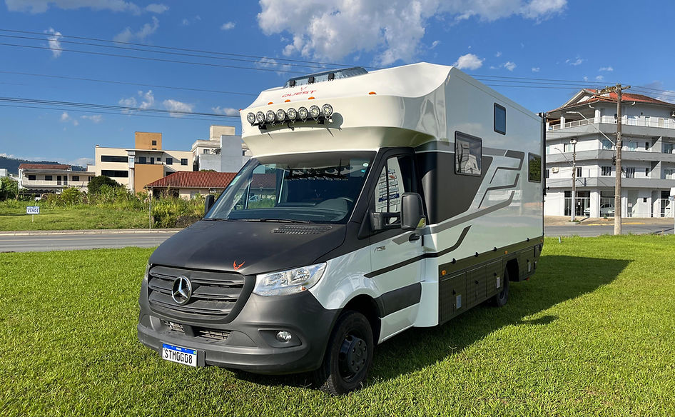 Motorhome estacionado em área arborizada com toldo aberto, mesa externa montad, transmitindo conforto.