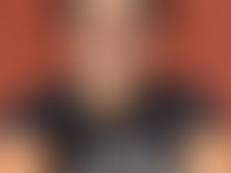 Smiling person with glasses in a black "Team Spirit" shirt, against a red wall. Sunny day, warm and cheerful vibe.