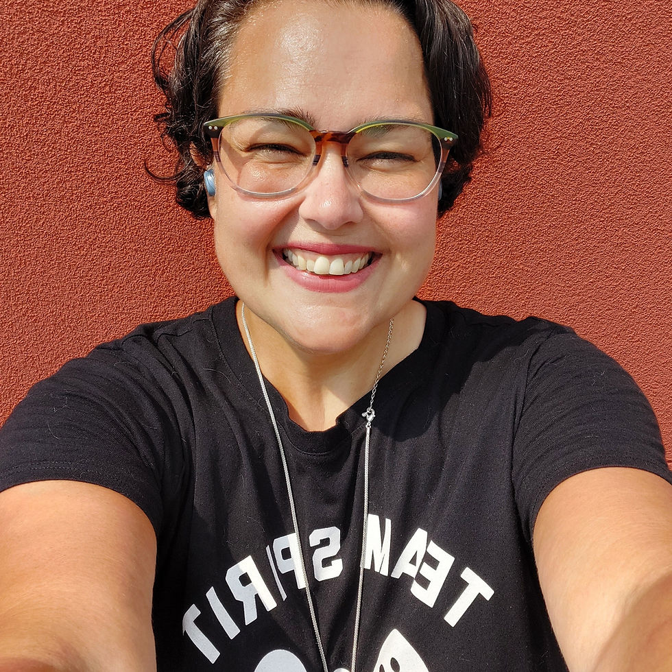 Smiling person with glasses in a black "Team Spirit" shirt, against a red wall. Sunny day, warm and cheerful vibe.