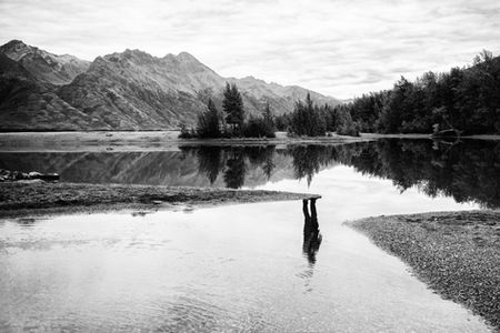 Black-and-white surreal landscape photograph of a calm mountain lake where a partially hidden human figure appears primarily through its dark reflection in the water.. Personal exhibition photograph