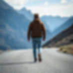 Person in a brown hoodie and jeans walks alone down a mountain road. Distant blue mountains and a clear sky create a serene backdrop.