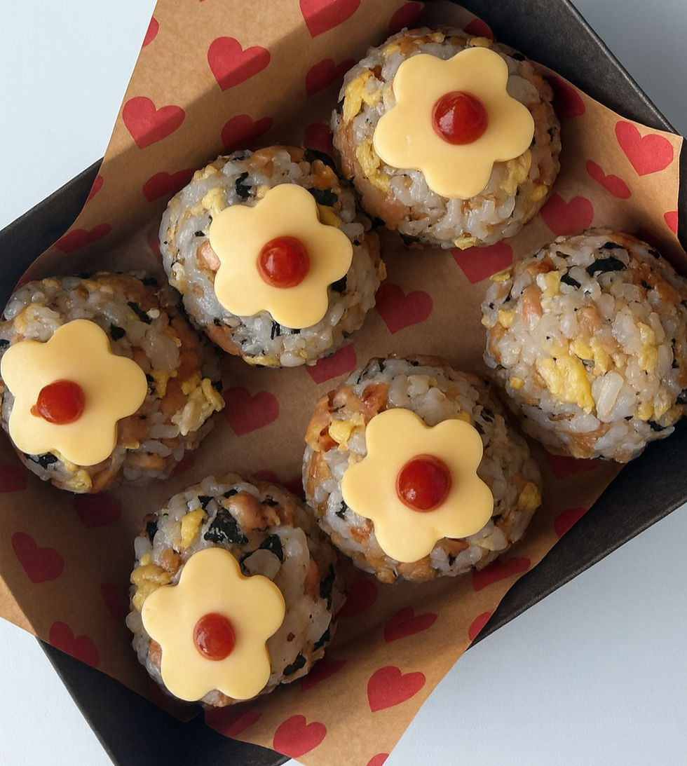 Six Korean rice balls topped with cheddar cheese flowers and ketchup dots, arranged in a heart-patterned paper-lined box.