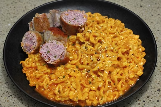Creamy spicy Korean ramen served on a black plate with sliced sausage pieces on top, garnished with sesame seeds on a kitchen countertop.