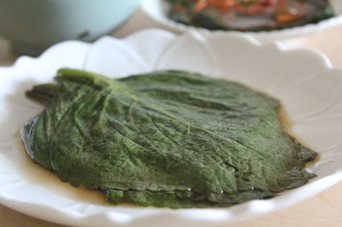 A large green leaf rests on a white scalloped plate with a little liquid around it, photographed close up on a light tabletop with another dish blurred in the background.