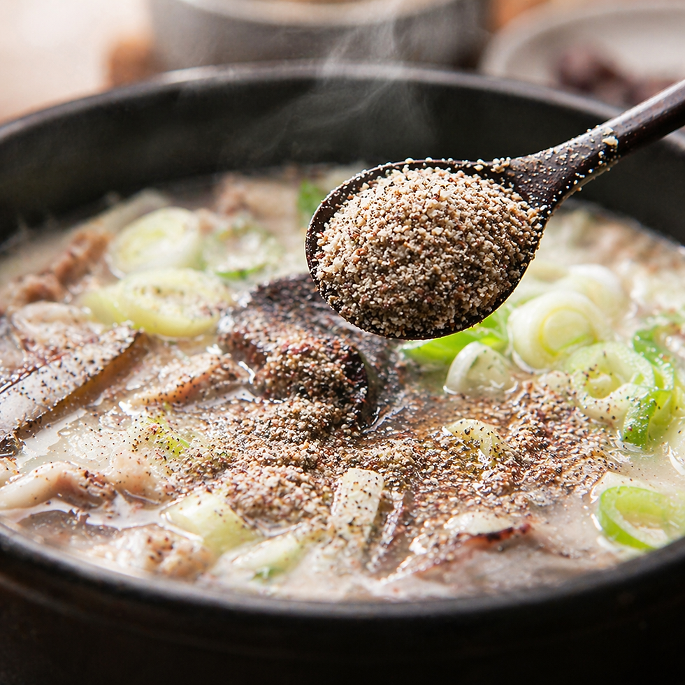 Close-up of a spoonful of ground deulkkae hovering over a steaming bowl of gukbap, with the nutty perilla seed powder falling into the broth among sliced green onions and meat.
