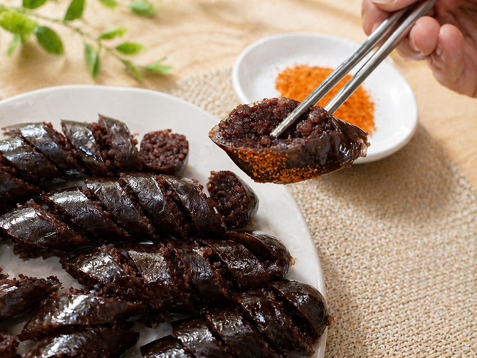 Metal chopsticks lift a slice of sundae above a plate of glossy sliced Korean blood sausage, with a small dish of orange dipping salt in the background on a warm, rustic tabletop.