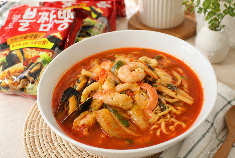 A white bowl of Paldo Bul Jjamppong spicy seafood noodle soup sits in a warm kitchen setting, filled with red broth, noodles, shrimp, mussels, squid, and vegetables, with the red Paldo Bul Jjamppong package beside it.