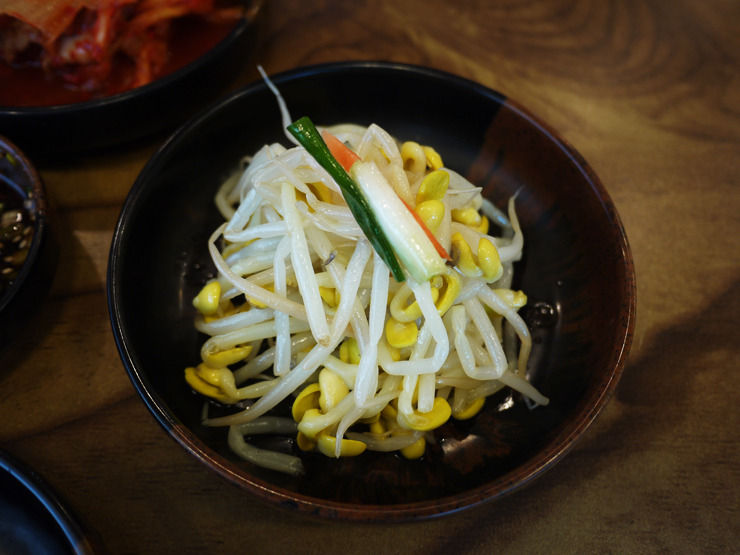 Korean seasoned soybean sprout banchan with scallion and red pepper garnish, served in a small black bowl on a wooden table.