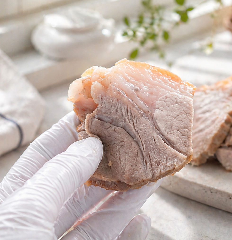 A gloved hand holding a tender slice of bossam pork, showing the moist meat and glossy fat layers with more sliced pork in the background.