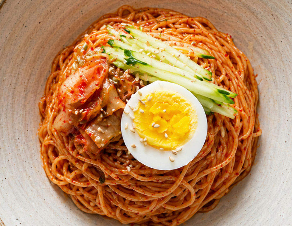 Close up shot of Bibim guksu noodles topped with cucumber strips, kimchi, sesame seeds, and half a boiled egg in a wide ceramic bowl on a dark stone surface.