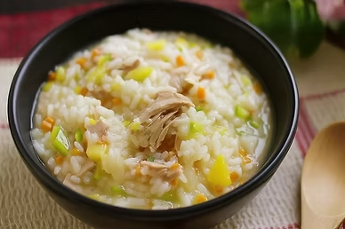 Chicken juk rice porridge with shredded chicken, vegetables, and soft rice served in a black bowl with a wooden spoon nearby.