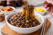 Chopsticks lifting jjajangmyeon noodles coated in black bean sauce above the bowl.